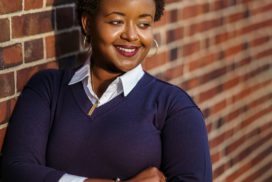 Smartly dressed Black woman smiling confidently, standing in front of a brick wall.