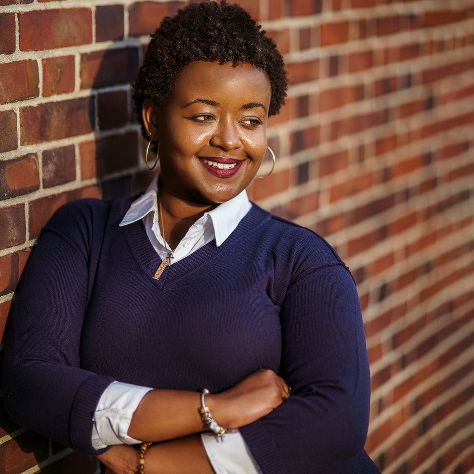 Smartly dressed Black woman smiling confidently, standing in front of a brick wall.