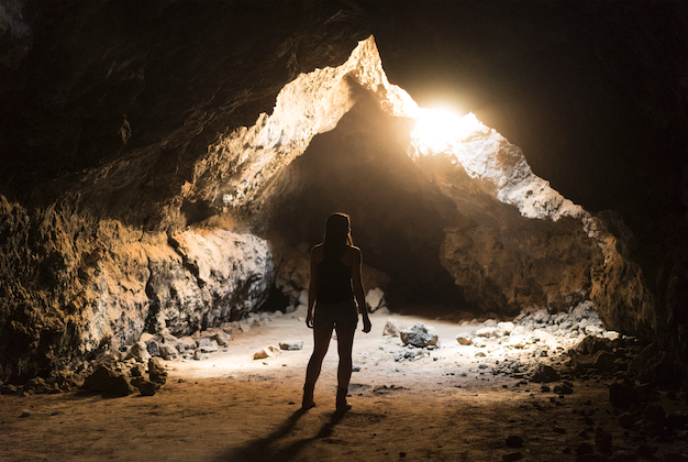 a woman enters a cave and regards the darkness in front of her. light streams in from above