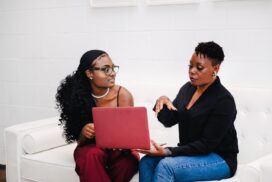 two professional women sit together on a white couch in a work meeting, looking at a laptop