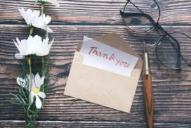 a wooden table top with white flowers, an interview thank you card and calligraphy pen, and a pair of glasses